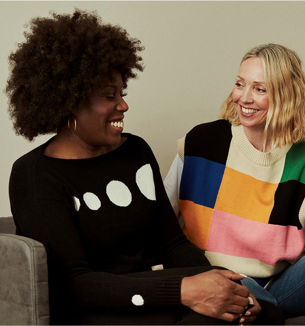 Deux femmes souriantes, assises côte à côte sur un canapé, engagées dans une conversation amicale. La femme à gauche porte un pull noir à motifs de cercles blancs, tandis que celle à droite porte un pull multicolore à motifs géométriques.